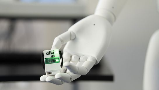 Roboterhand hält Computerchip mit DFKI-Logo Beschriftung