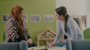 Das Bild zeigt zwei Frauen, die sich gegenüber sitzen. Zwischen ihnen steht ein Tisch auf dem ein Tablet liegt. Die eine Frau trägt ein Kopftuch. Die andere Frau hat braune Haare und trägt einen Zopf. Die Frau mit dem Zopf hat ihre Hand auf dem Tablet. Die Frauen lächeln sich an. Im Hintergrund sieht man eine grüne Wand und ein Puppenhaus.