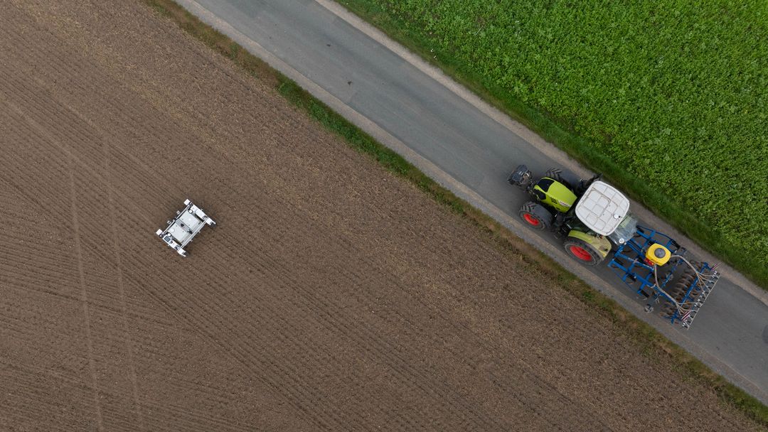 Aus der Vogelperspektive: Ein Roboter auf einem Feld, ein Weg, auf dem ein Traktor fährt.