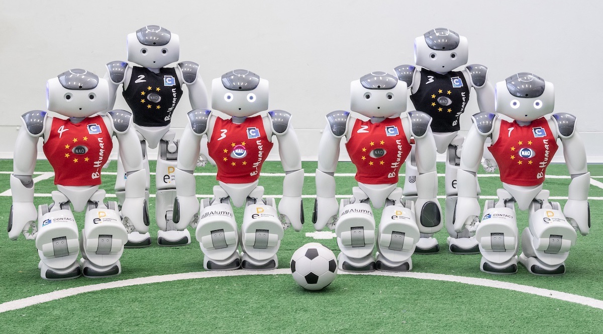 Teambild: sechs Roboter stehen aufgereiht an der Spielfeldlinie, Frontal zur Kamera, vor ihnen der Ball.