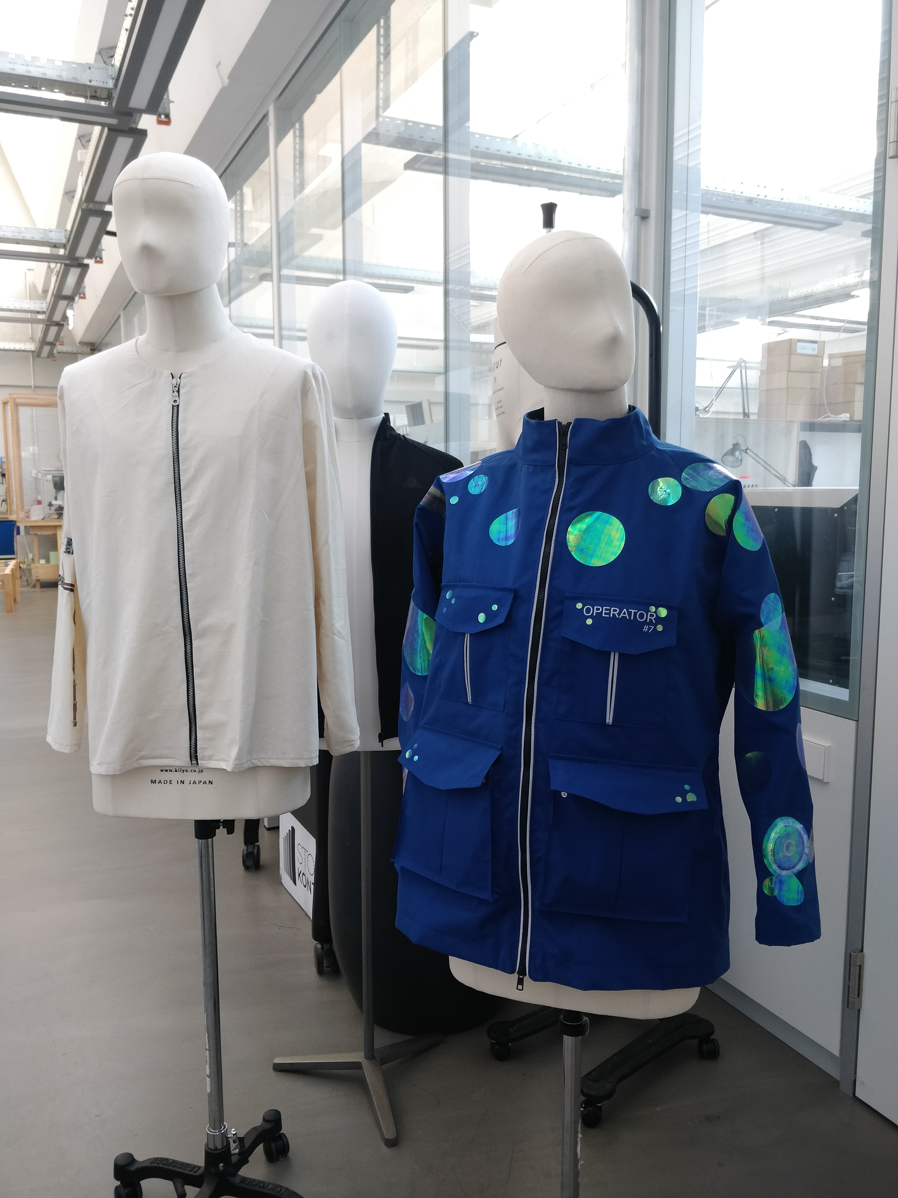 Das Bild zeigt zwei Schneiderpuppen, die Prototypen von Jacken tragen. Die linke Figur trägt eine weißte Jacken, während die rechte Figur eine blaue Jacke trägt. Die blaue Jacke ist ausgestattet mit Sensoren und reflektierenden Patches. 